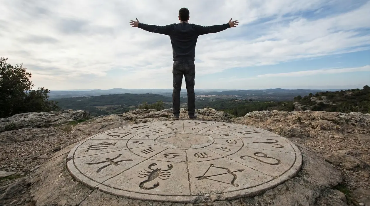 Persona su una piattaforma con simboli zodiacali scolpiti, fronte a un paesaggio montano