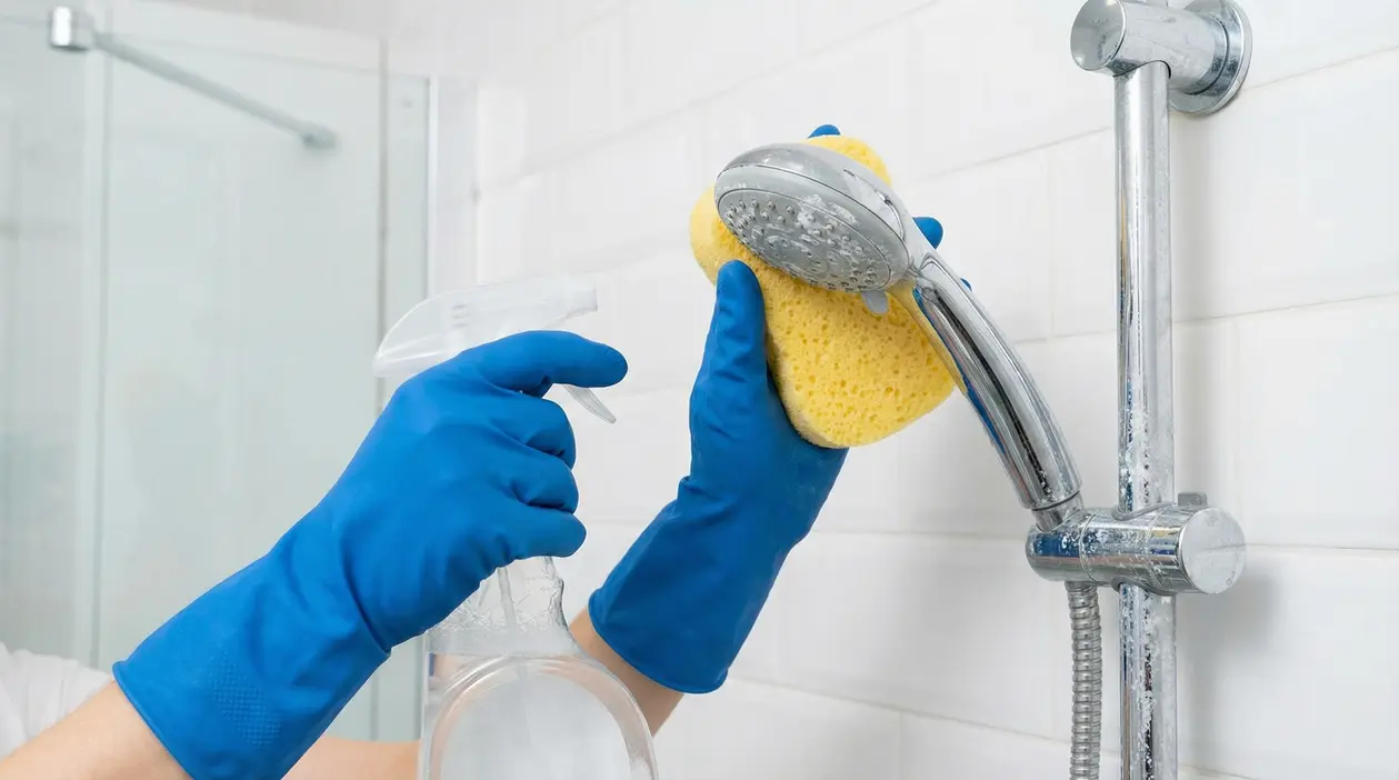 Una persona pulisce un soffione della doccia con una spugna e un flacone spray